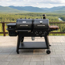 A BBQ Boards® Pit Boss Pellet Gas Combo Side Cutting Board sits on a stone patio next to a grill and smoker, overlooking dense green forest and mountains, providing convenient prep space.