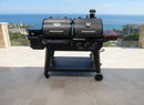 A BBQ Boards® Pit Boss Pellet Gas Combo Side Cutting Board by BBQ Boards® is attached to a large black grill with two compartments, set on a tiled patio overlooking the ocean and coastal buildings on a sunny day.