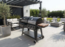 A modern patio features a black grill with the BBQ Boards® Pit Boss Pellet Gas Combo Side Cutting Board by BBQ Boards®, surrounded by white cushioned seating, potted plants, a pergola, and set on light decking overlooking a green yard.