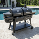 A BBQ Boards® Pit Boss Pellet Gas Combo Side Cutting Board is attached to a two-compartment grill with control knobs, set on a tiled patio beside a pool, surrounded by greenery and a modern house.