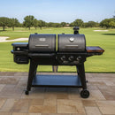 A BBQ Boards® Pit Boss Pellet Gas Combo Side Cutting Board is attached to a black combo grill with dual lids and a side burner, positioned on a patio that overlooks a lush golf course lined with trees.