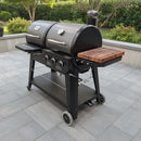 A black Pit Boss Pellet Gas Combo grill by BBQ Boards® features silver handles, control knobs, a built-in thermometer, and a BBQ Boards® Side Cutting Board attached on the right. Green bushes can be seen in the background.