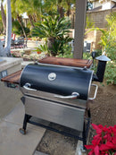 A sunny backyard patio features the BBQ Boards® Recteq RT-700 Pair, Front & Side Cutting Boards by BBQ Boards®, installed on a black and stainless steel outdoor cooking station surrounded by lush greenery.