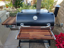 The BBQ Boards® Recteq RT-700 Pair, Front & Side Cutting Boards by BBQ Boards® are set up on a patio beside a garden and brick wall, creating an ideal outdoor cooking station near red flowers.
