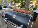 A black outdoor cooking station with stainless steel handles stands on a patio. BBQ Boards® Recteq RT-700 Front & Side Cutting Boards (Pair) are on the side shelves. Palm trees, patio furniture, and a pool surrounded by greenery are in the background.
