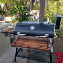 On a sunny patio with greenery and furniture, BBQ Boards® Recteq RT-700 Front & Side Cutting Boards (Pair) by BBQ Boards® are set on a black and stainless grill with a wooden shelf for convenient BBQ prep.