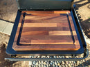The BBQ Boards® Traeger Flatrock Side Cutting Board sits on a Traeger Flatrock side board, displaying the BBQ Boards® logo and providing a convenient prep space above visible stones and dirt.