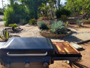 A BBQ Boards® Traeger Flatrock Side Cutting Board, by BBQ Boards®, doubles as a prep and serving shelf on a Traeger grill set in a sunlit garden with agave, stone pathways, and lush greenery.