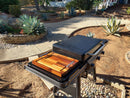 A BBQ Boards® Traeger Flatrock Side Cutting Board by BBQ Boards® is shown on a closed black grill with a wooden surface, set on a stone patio in a landscaped backyard with agave, gravel, and stone pathway. Sunlight casts shadows over the scene.
