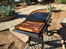 A gas grill with a closed lid stands on an outdoor pebble area beside a BBQ Boards® Traeger Flatrock Side Cutting Board, providing prep space. Around the grill are stone-edged garden beds with agave and paved walkways.