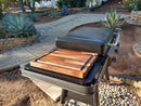 A BBQ Boards® Traeger Flatrock Side Cutting Board by BBQ Boards® provides a convenient cutting and serving surface on a grill with a closed lid, set on a patio with stone paths, agave plants, desert landscaping, and steps in the background.