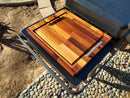 The BBQ Boards® Traeger Flatrock Side Cutting Board by BBQ Boards® adds extra BBQ prep space outdoors, resting on the grill’s side board near a stone walkway and dry soil, and features a large wooden surface with metal handle.