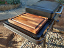 The BBQ Boards® Traeger Flatrock Side Cutting Board adds a wooden cutting surface to your black outdoor grill, set against a stone walkway, lush plants, and a raised garden bed.