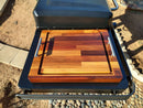 The BBQ Boards® Traeger Flatrock Side Cutting Board by BBQ Boards® rests on an outdoor grill’s side shelf, providing a spacious wooden prep area with metal handles, against a backdrop of dirt, stones, and paving.