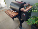 The BBQ Boards® Traeger Ironwood 650 Front & Side Cutting Boards (Pair) by BBQ Boards® create a perfect patio BBQ workspace. Three seasoning jars and a green potted plant sit on the side board near a light gray outdoor wall.