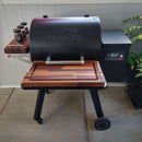 A black Traeger Ironwood 650 grill with BBQ Boards® Front & Side Cutting Boards (Pair) creates a BBQ prep station with wood shelves and spice storage. Digital controls, pellet hopper, and green plants to the right complete the patio scene.