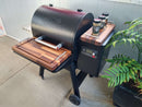 The BBQ Boards® Traeger Ironwood 650 Front & Side Cutting Boards (Pair) by BBQ Boards® enhance your grilling setup with extra prep space for spices and utensils, fitting seamlessly onto your Traeger grill on a concrete patio beside a green potted plant.