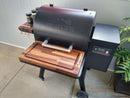 A Traeger Ironwood 650 grill with BBQ Boards® Front & Side Cutting Boards creates an ideal BBQ setup, with spice jars on the side rack, digital controls on the right, and a potted plant—set on concrete by a light-colored wall.