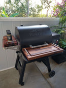 A Traeger Ironwood 650 grill on a patio features BBQ Boards® Traeger Ironwood 650 Front & Side Cutting Boards (Pair) as spacious work surfaces, with spice jars on the side shelf and plants and trees in the background.