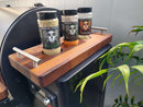 Three spice jars labeled Smaklish Asada Rub sit on a wooden tray with metal handles, placed on an outdoor grill’s side shelf. A green plant is nearby, and BBQ Boards® Traeger Ironwood 650 Front & Side Cutting Boards accent the BBQ workspace.