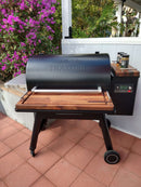 A black Traeger pellet grill with BBQ Boards® Traeger Ironwood 885 Front & Side Cutting Boards (Pair) stands on a tiled patio by pink flowers and a white fence, with seasonings and grilling accessories on the prep station.