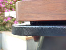 Close-up side view of a dark metal and wood surface, likely a BBQ Boards® Traeger Ironwood 885 Front & Side Cutting Board (Pair) by BBQ Boards®, with a blurred background featuring pink flowers and greenery.