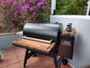 A Traeger Ironwood 885 with BBQ Boards® Front & Side Cutting Boards (Pair) sits on a tiled patio by purple flowers. Two spice containers rest on the BBQ Boards® sideboards, perfect for convenient grilling prep.
