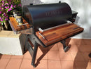 A black Traeger Ironwood 885 grill is fitted with BBQ Boards® Front & Side Cutting Boards, used as a BBQ prep station. Several seasoning jars sit on the boards, with nearby flowers and sunlight on a tiled patio.