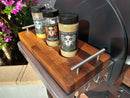 Three “Spanglish Asadero” spice bottles sit on a BBQ Boards® Traeger Ironwood 885 Front & Side Cutting Board, which has metal handles. Purple flowers are visible in the background.