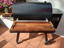 A Traeger pellet grill fitted with BBQ Boards® Traeger Ironwood 885 Front & Side Cutting Boards (Pair) stands on a patio, three seasoning jars on its side shelf. Pink flowers bloom by the white fence, making an ideal BBQ prep station.