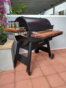 A black Traeger Ironwood 885 with BBQ Boards® Front & Side Cutting Boards (Pair) stands on a red-tiled patio. Three spice jars sit on the left board, with purple flowers visible to the left of the grill.