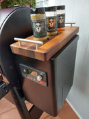 Three spice jars with vibrant skull labels rest on BBQ Boards® Traeger Ironwood 885 Front & Side Cutting Boards (Pair) by BBQ Boards®, positioned beside the control panel of a black grill, creating an ideal BBQ prep area.