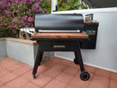 A black Traeger Ironwood 885 pellet grill with BBQ Boards® Traeger Ironwood 885 Front & Side Cutting Boards (Pair) stands on a red-tiled patio by a gray wall and flowering bushes, featuring the grill's control panel on the right.