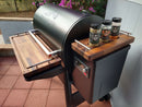 A BBQ Boards® Traeger Ironwood 885 Front & Side Cutting Boards (Pair) set is installed on a Traeger grill, surrounded by blooming flowers and a white fence—a stylish BBQ prep station with seasoning jars and a digital control panel.