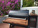 The BBQ Boards® Traeger Ironwood 885 Front & Side Cutting Boards (Pair) by BBQ Boards® are displayed on a Traeger grill with a wooden shelf on a patio, surrounded by blooming purple flowers and greenery, grilling accessories nearby.