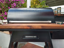A Traeger Ironwood 885 pellet grill with a closed lid is shown outdoors, featuring BBQ Boards® Traeger Ironwood 885 Front & Side Cutting Boards (Pair) as shelves. Flowering bushes and seasoning cans complete the ideal BBQ prep station scene.