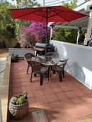 A patio with a red umbrella shades a glass table and four plastic chairs. BBQ Boards® Traeger Ironwood 885 Front & Side Cutting Boards (Pair) by BBQ Boards® are on display near the grill, with potted cacti and purple flowers adding color to the tiled area.