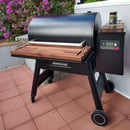 A Traeger Ironwood 885 grill is fitted with BBQ Boards® Front & Side Cutting Boards (Pair) and stands on a red-tiled patio near purple flowers, featuring a wood shelf, control panel, and a drink on the BBQ Boards® prep station.