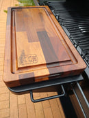 The BBQ Boards® Traeger Ironwood Front Cutting Board sits on the side shelf of a black outdoor grill, serving as a clean and convenient BBQ prep station on the brick patio.