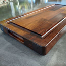 The BBQ Boards® Traeger Ironwood Front Cutting Board features a polished wood surface with rounded edges, a built-in side handle, and sits on a gray countertop—ideal for any BBQ prep station.