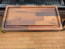 The BBQ Boards® Traeger Ironwood XL Front Cutting Board features a rectangular wooden design with an edge groove and BBQ Boards® logo, displayed atop the Traeger Ironwood XL front shelf above a brick patio.