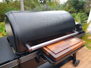 A black Traeger grill with a closed lid sits on the patio. Its shelf features the BBQ Boards® Traeger Ironwood XL Front Cutting Board by BBQ Boards®, perfect for BBQ prep, set against lush green plants and trees.