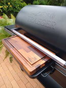 A close-up of a BBQ Boards® Traeger Ironwood XL Front Cutting Board by BBQ Boards® on a Traeger grill’s side shelf, creating an ideal BBQ prep space, with greenery and brick patio flooring in the background.