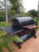 A large, black Traeger grill with a BBQ Boards® Traeger Ironwood XL Front Cutting Board stands on a brick patio surrounded by lush greenery, flowers, and trees—the perfect BBQ prep space in a beautiful garden setting.