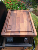 The BBQ Boards® Traeger Ironwood XL Pellet Bin Cutting Board, featuring a corner BBQ Boards® logo and an edge groove, sits atop a Traeger Ironwood XL surrounded by plants and flowers.