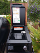 An open BBQ Boards® Traeger Ironwood XL Pellet Bin Cutting Board by BBQ Boards® is shown outdoors among plants and flowers, revealing the inside compartment, control panel, and safety instructions on the lid. A digital probe rests on the cutting surface.