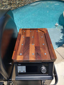 A BBQ Boards® Traeger Ironwood XL Pellet Bin Cutting Board by BBQ Boards® sits atop the grill, serving as a cutting and staging surface beside a pool with clear blue water and stone edging in the background.