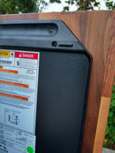 Close-up of a BBQ Boards® Traeger Ironwood XL Pellet Bin Cutting Board by BBQ Boards®, showing warning labels and a black plastic panel with screws against simulated wood grain. Green plants and trees are blurred in the background.