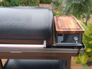 A close-up of a BBQ Boards® Traeger Ironwood XL Pellet Bin Cutting Board being used as a cutting and staging surface on the grill’s side shelf, with the digital control panel and plants in the background.