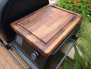 The BBQ Boards® Traeger Ironwood XL Pellet Bin Cutting Board sits on a black grill side shelf, framed by green ferns and red flowers—an ideal wooden prep surface for your Traeger Ironwood XL.